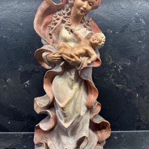 Könnte beinhalten: Detaillierte Statue, die eine Frau mit einem Baby zeigt. Die Statue ist in gedämpften Beige-, Rosa- und Brauntönen bemalt. Die Frau trägt ein drapiertes Gewand und eine Kopfbedeckung. Das Baby wird in ihren Armen gehalten. Die Statue steht auf einem runden Sockel.