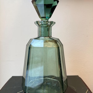 Könnte beinhalten: Eine grüne Glasdekanter mit einem facettierten, rautenförmigen Verschluss. Die Flasche hat ein geometrisches Design und eine breite Basis, die sich nach oben verjüngt. Der Verschluss hat einen dunkleren Grünton als die Flasche.