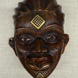 Könnte beinhalten: Detaillierte, braune Holzmaske mit aufwendigen Schnitzereien und einem stilisierten Gesicht. Die Maske hat eine markante Stirn, detaillierte Augen und einen lächelnden Mund. Goldfarbene rautenförmige Akzente befinden sich auf Stirn und Kinn. Ein kulturelles Artefakt.