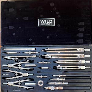 以下が含まれることがあります： 黒いケースには、Wild Heerbrugg 製の製図ツール一式が入っています。このセットには、さまざまなコンパス、ディバイダー、その他の精密機器が含まれており、すべてシルバーと黒です。ケースには、ブランド名「WILD HEERBRUGG」が白い長方形で表示されています。