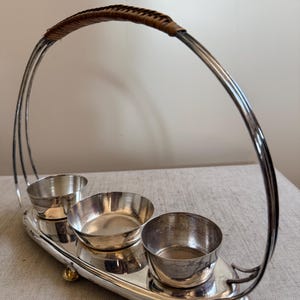 Könnte beinhalten: Silberfarbenes Serviertablett mit drei kleinen Schalen. Das Tablett hat einen gebogenen Griff mit einem geflochtenen braunen Griff. Die Schalen sind rund und stehen auf einer ovalen Basis. Das Tablett hat kleine goldfarbene Füße.