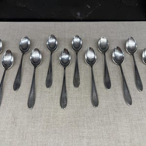 May include: A collection of twelve silver spoons arranged on a light beige surface. The spoons have a classic design with oval bowls and long, slender handles. The spoons are evenly spaced and reflect light.