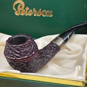 Peut inclure: Une pipe à tabac Peterson avec un foyer texturé rouge foncé et un tuyau noir. La pipe est présentée dans un coffret vert avec le nom de la marque "Peterson" en or sur le couvercle. L'intérieur de la boîte est doublé de tissu crème.