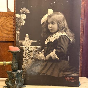 Puede incluir: Fotografía sepia de una niña con vestido oscuro, cuello y lazo de encaje, fechada en 1912. Un candelabro decorativo y un jarrón con flores en primer plano. Retrato infantil de estilo vintage.