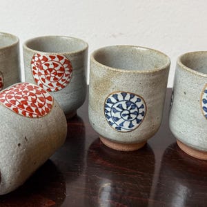 Könnte beinhalten: Set aus fünf Keramikbechern mit gesprenkelter grauer Oberfläche. Jeder Becher hat ein kreisförmiges Design in Rot oder Blau. Die Becher haben ein rustikales, handgefertigtes Aussehen mit braunem Boden.