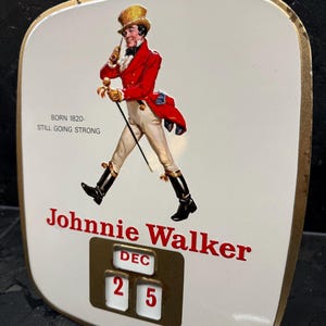 Puede incluir: Letrero vintage de Johnnie Walker con fondo blanco y borde dorado. Presenta un hombre con abrigo rojo y sombrero de copa, el texto "Johnnie Walker" y la fecha "25 DIC". El letrero tambi&eacute;n incluye el texto "BORN 1820 - STILL GOING STRONG".