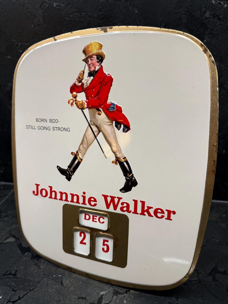 Puede incluir: Letrero vintage de Johnnie Walker con fondo blanco y borde dorado. Presenta un hombre con abrigo rojo y sombrero de copa, el texto "Johnnie Walker" y la fecha "25 DIC". El letrero tambi&eacute;n incluye el texto "BORN 1820 - STILL GOING STRONG".