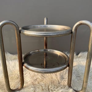 Könnte beinhalten: Ein zweistufiger Beistelltisch aus Metall mit runden Ablagen. Der Tisch hat ein Vintage- und Industrie-Aussehen mit einer verwitterten, silbernen Oberfläche. Die gebogenen Beine und der Griff tragen zu seinem einzigartigen Design bei. Höhe ca. 40 cm.