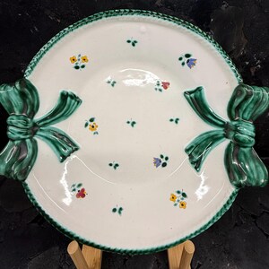 Könnte beinhalten: Eine weiße Keramik-Servierplatte mit einem grünen Schleifendesign auf jeder Seite. Die Platte hat einen gewellten Rand und ist mit kleinen floralen Akzenten in Gelb, Rot und Blau verziert. Die Platte hat einen Durchmesser von etwa 30 cm.