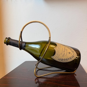 May include: A green champagne bottle in a gold-coloured metal wine holder with a woven handle. The bottle has a label that reads "Dom Pérignon Champagne Vintage 2006". The holder is on a dark wooden surface.