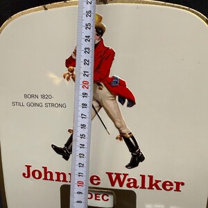 Puede incluir: Letrero vintage de Johnnie Walker con el ic&oacute;nico Striding Man con abrigo rojo, pantalones blancos y botas negras. El letrero es blanco con letras rojas e incluye el texto "BORN 1820 - STILL GOING STRONG". El calendario muestra "DEC 5". Mide aprox. 28 cm.