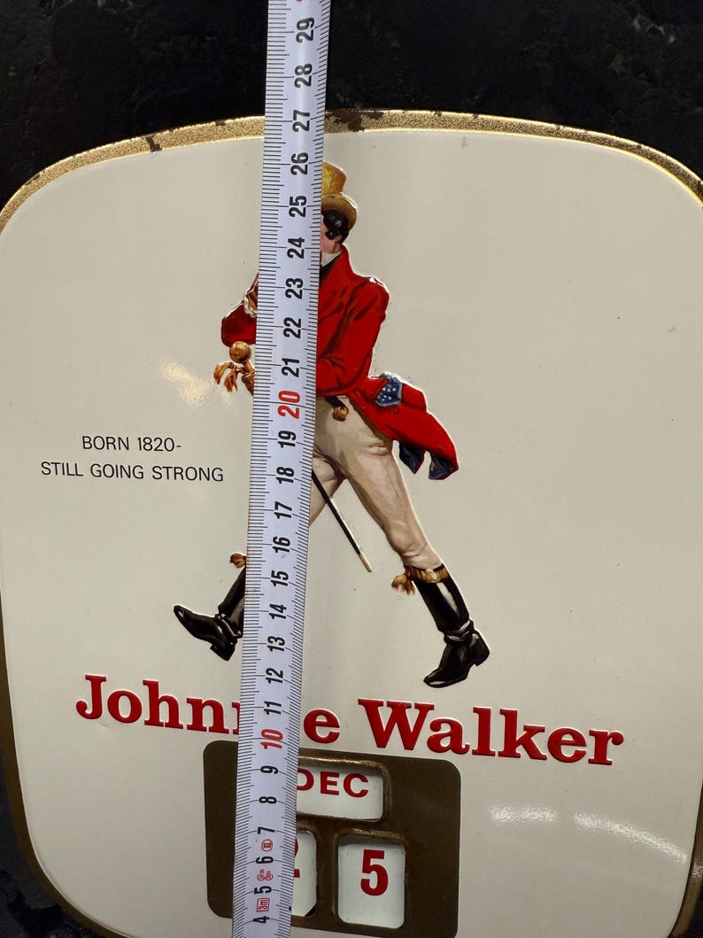 Puede incluir: Letrero vintage de Johnnie Walker con el ic&oacute;nico Striding Man con abrigo rojo, pantalones blancos y botas negras. El letrero es blanco con letras rojas e incluye el texto "BORN 1820 - STILL GOING STRONG". El calendario muestra "DEC 5". Mide aprox. 28 cm.