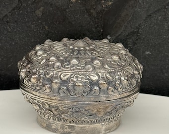 Op de afbeelding: Een zilverkleurige, sierlijke, ronde doos met een deksel. De doos heeft gedetailleerde bloemen- en stippenpatronen, met een licht verhoogd, getextureerd oppervlak. De basis is breder dan het deksel, waardoor een kenmerkende vorm ontstaat.