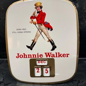 Puede incluir: Letrero publicitario vintage de Johnnie Walker con fondo blanco y borde dorado. El letrero presenta una figura con abrigo rojo con bast&oacute;n y el texto "Johnnie Walker". La fecha est&aacute; fijada en el 25 de diciembre.