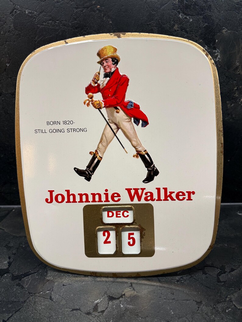 Puede incluir: Letrero publicitario vintage de Johnnie Walker con fondo blanco y borde dorado. El letrero presenta una figura con abrigo rojo con bast&oacute;n y el texto "Johnnie Walker". La fecha est&aacute; fijada en el 25 de diciembre.