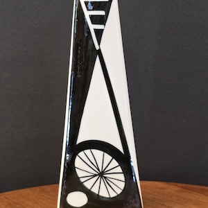 May include: A tall, triangular ceramic vase with a black and white geometric design. The vase features a black wheel-like shape and a white oval. The vase is on a wooden surface.