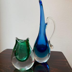 Puede incluir: Dos jarrones de cristal. Uno tiene forma de lágrima en cristal transparente con interior verde. El otro es una jarra alta y delgada de cristal azul con asa transparente. Ambos están sobre una superficie de madera oscura.