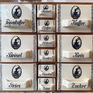 Puede incluir: Un especiero vintage con cajones de cerámica blanca y marco de madera. Cada cajón está etiquetado con un nombre de especia diferente en escritura negra. Los cajones presentan un diseño ovalado decorativo en negro y azul con una mujer sosteniendo un paraguas.
