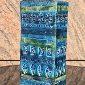 May include: A rectangular, blue ceramic vase with a textured surface and geometric patterns. The vase features a mix of blue and green hues, with decorative elements including squares, diamonds, and lines. The vase is approximately 20 cm tall.