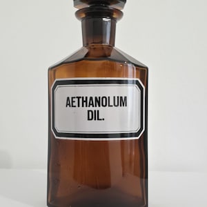 Könnte beinhalten: Eine braune Apothekerflasche aus Glas mit einem schwarzen Verschluss. Die Flasche hat ein weißes Etikett mit schwarzem Text, auf dem "AETHANOLUM DIL." steht. Die Flasche ist rechteckig mit breitem Boden und schmalem Hals.