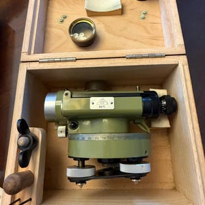 May include: An open wooden box containing a green and silver surveying level with a circular base and a black eyepiece. The box also holds small accessories, including a small round container and a wooden handle. The level has the number "8071" on it.