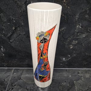 Könnte beinhalten: Eine hohe, cremefarbene Keramikvase mit vertikaler Streifenstruktur. Die Vase zeigt eine farbenfrohe Illustration einer Frau in blauem Kleid und rotem Schal, die eine Blume hält. Die Vase ist etwa 25 cm hoch.