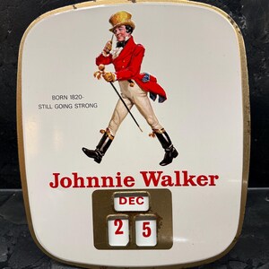 Puede incluir: Art&iacute;culo publicitario vintage de Johnnie Walker con fondo blanco y borde dorado. La imagen presenta a un hombre con abrigo rojo y el texto "Johnnie Walker". La fecha est&aacute; fijada al 25 de diciembre. El texto dice "Nacido en 1820 - Todav&iacute;a fuerte."