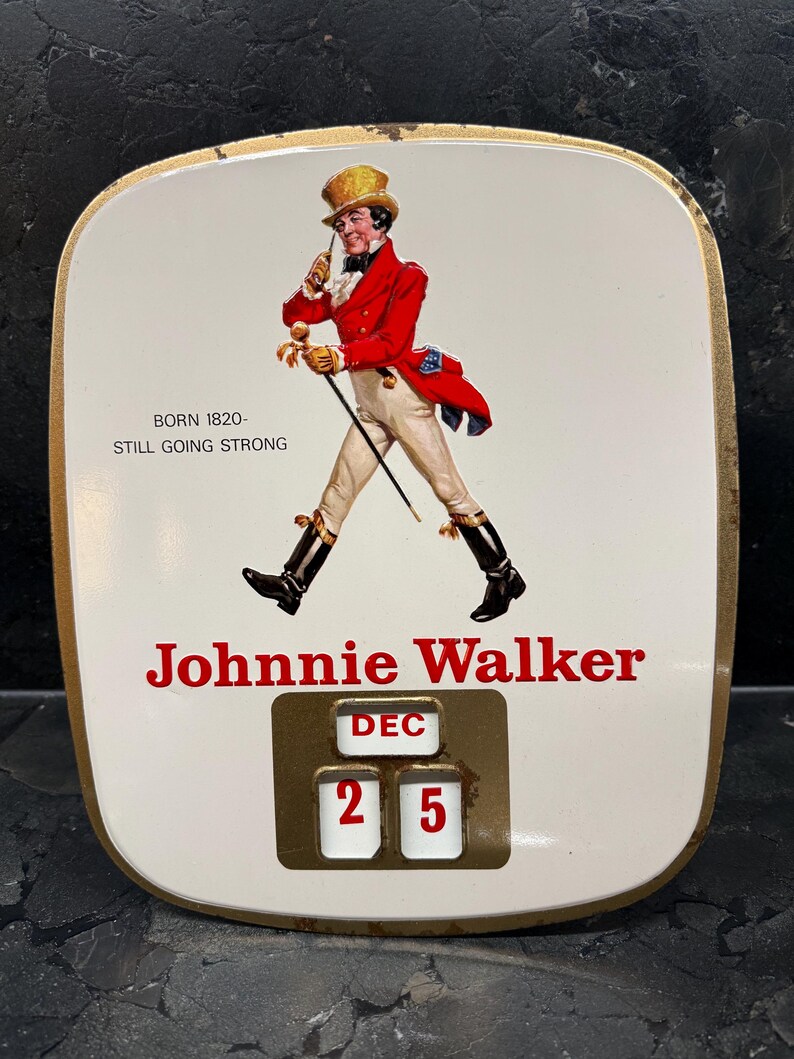 Puede incluir: Art&iacute;culo publicitario vintage de Johnnie Walker con fondo blanco y borde dorado. La imagen presenta a un hombre con abrigo rojo y el texto "Johnnie Walker". La fecha est&aacute; fijada al 25 de diciembre. El texto dice "Nacido en 1820 - Todav&iacute;a fuerte."