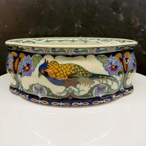 May include: A decorative ceramic bowl with a pheasant design. The bowl features a cream-colored base with a detailed illustration of a pheasant in shades of gold, blue, and brown. Floral patterns in blue and purple adorn the sides, with a dark blue border.