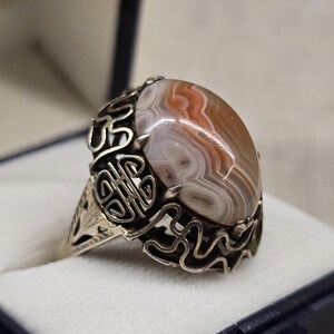 Könnte beinhalten: Ein Silberring mit einem ovalen Achatstein. Der Stein zeigt eine Mischung aus weißen, braunen und orangefarbenen Farbtönen. Der Ring hat ein verziertes, durchbrochenes Design um den Stein. Der Ring befindet sich in einer dunkelblauen Schachtel.