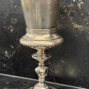 Könnte beinhalten: Ein silberfarbener Kelch mit einem breiten, zylindrischen Oberteil und einem dekorativen Sockel. Der Kelch hat ein detailliertes, verziertes Design auf dem Sockel und dem Stiel, mit einer glatten, polierten Oberfläche auf der Tasse. Das Gesamtdesign ist vintage und elegant.