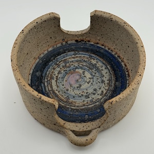 Könnte beinhalten: Eine runde, gesprenkelte beige Keramik-Garnschale mit glattem, glänzendem Innenraum und einem blaugrauen Wirbelmuster. Die Schale hat eine tiefe U-förmige Aussparung am Rand für den Garnfaden und einen kleinen, kreisförmigen Griff an der Basis.