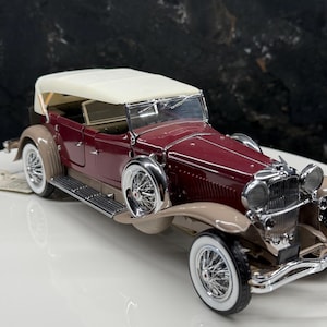 May include: A detailed model of a classic burgundy convertible car with a cream-colored top. The car features chrome accents, white-walled tires, and tan fenders. The model is a vintage replica, showcasing intricate design details.