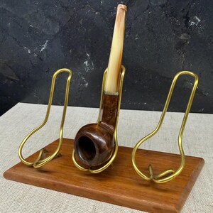 Könnte beinhalten: Eine Holzpfeife mit hellem Mundstück ruht in einem Messinghalter auf einem Holzsockel. Der Halter hat ein gebogenes Design. Die Pfeife ist dunkelbraun mit einem abgerundeten Kopf. Der Sockel ist rechteckig und aus warm getöntem Holz.