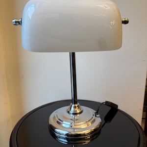 Könnte beinhalten: Eine weiße Banker-Lampe mit poliertem Chromfuß und -schaft. Die Lampe hat einen abgerundeten, undurchsichtigen weißen Schirm und kleine, dekorative Metallakzente auf jeder Seite. Die Lampe steht auf einem schwarzen, glänzenden Tisch.