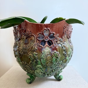 May include: A decorative ceramic planter with a detailed floral and scrollwork design. The planter features a rich color palette of green, brown, and bronze. It has three small feet and a flower-shaped cutout. The planter is filled with green leaves.