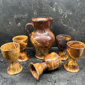 Könnte beinhalten: Ein handgefertigter Keramikkrug und sechs passende Becher. Das Set hat eine warme braune Glasur mit helleren, marmorierten Akzenten. Der Krug hat einen Henkel und einen Ausguss. Die Becher haben einen Sockel.