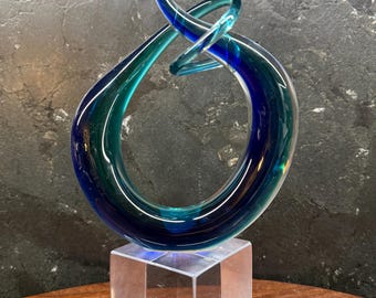 May include: A decorative glass sculpture featuring a swirling, teardrop-shaped form in shades of deep blue and teal. The glass piece is mounted on a clear, transparent, square-shaped glass base, creating a smooth, reflective, and polished appearance.