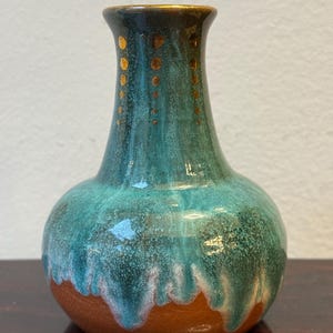 Könnte beinhalten: Eine Keramikvase mit bauchigem Körper und schmalem Hals. Die Vase hat eine türkis-braune Glasur mit Tropfeffekt. Der Rand ist dunkelbraun mit goldenen Punkten. Die Vase ist etwa 15 cm hoch.