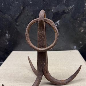 Könnte beinhalten: Ein rostiger, antiker Metallhaken mit einem kreisförmigen Oberteil und drei gebogenen Zinken. Der Haken besteht aus Eisen und weist ein verwittertes Aussehen auf, das auf Alter und Gebrauch hindeutet. Das Design ist einfach und funktional, wahrscheinlich zum Aufhängen oder Halten von Gegenständen.