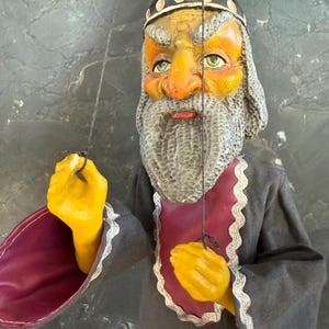 Könnte beinhalten: Eine Vintage-Puppe eines bärtigen Mannes mit Krone und langem grauen Bart. Die Puppe hat ein gelbes Gesicht und gelbe Hände sowie eine rote und graue Robe. Die Puppe hält Fäden, um ihre Bewegungen zu steuern.