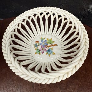 Könnte beinhalten: Ein weißer Keramikkorb mit einem gewebten Design. Der Korb hat ein zentrales Blumenmotiv mit rosa, blauen und gelben Blumen und grünen Blättern. Der Rand hat ein geflochtenes Seilmuster.