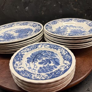 Könnte beinhalten: Eine Sammlung von blauen und weißen Porzellantellern im Vintage-Stil. Jeder Teller zeigt ein detailliertes Landschaftsdesign mit floralen Rändern. Die Teller sind in drei Stapeln auf einer Holzoberfläche gestapelt und bieten eine klassische Ästhetik.