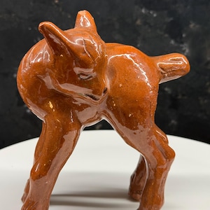 Könnte beinhalten: Glasierte Keramikfigur eines Hirsches in stehender Position, Kopf zur Seite gedreht. Der Hirsch ist warmbraun mit glänzendem Finish. Die Figur hat detaillierte Beine und einen kleinen Schwanz und zeigt eine handwerkliche Ästhetik.