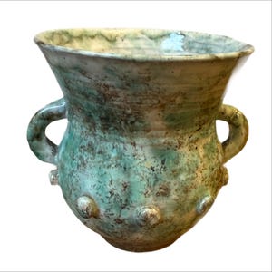 Könnte beinhalten: Eine handgefertigte Keramikvase mit breitem, ausgestelltem Rand und zwei Schlaufengriffen. Die Vase hat eine gesprenkelte türkisfarbene und braune Glasur, eine strukturierte Oberfläche und dekorative Knöpfe am Boden. Geeignet für die Inneneinrichtung.
