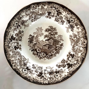 May include: A brown and white decorative plate with a floral and landscape design. The plate features a central scene of a house and trees, surrounded by a border of flowers and scrollwork. The rim is a darker brown color.