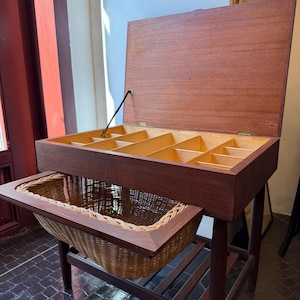 Op de afbeelding: Een vintage houten opbergkast met een opklapbaar deksel en verdeelde compartimenten. Onder de kast bevindt zich een uittrekbare geweven mand. De kast wordt ondersteund door vier poten en een onderste plank. Het hout heeft een warme, roodbruine tint.
