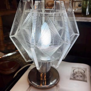 Op de afbeelding: Een heldere, geometrische tafellamp met een uniek ontwerp. De lamp heeft een heldere, veelzijdige kap met een webachtig patroon, ondersteund door een glanzende, zilverkleurige voet. De gloeilamp is zichtbaar aan de binnenkant.