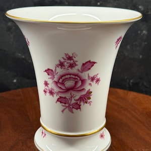 May include: White porcelain vase with a flared top and a footed base, adorned with a pink floral design. The vase features gold trim along the rim and base, adding a touch of elegance. A decorative piece for home decor.