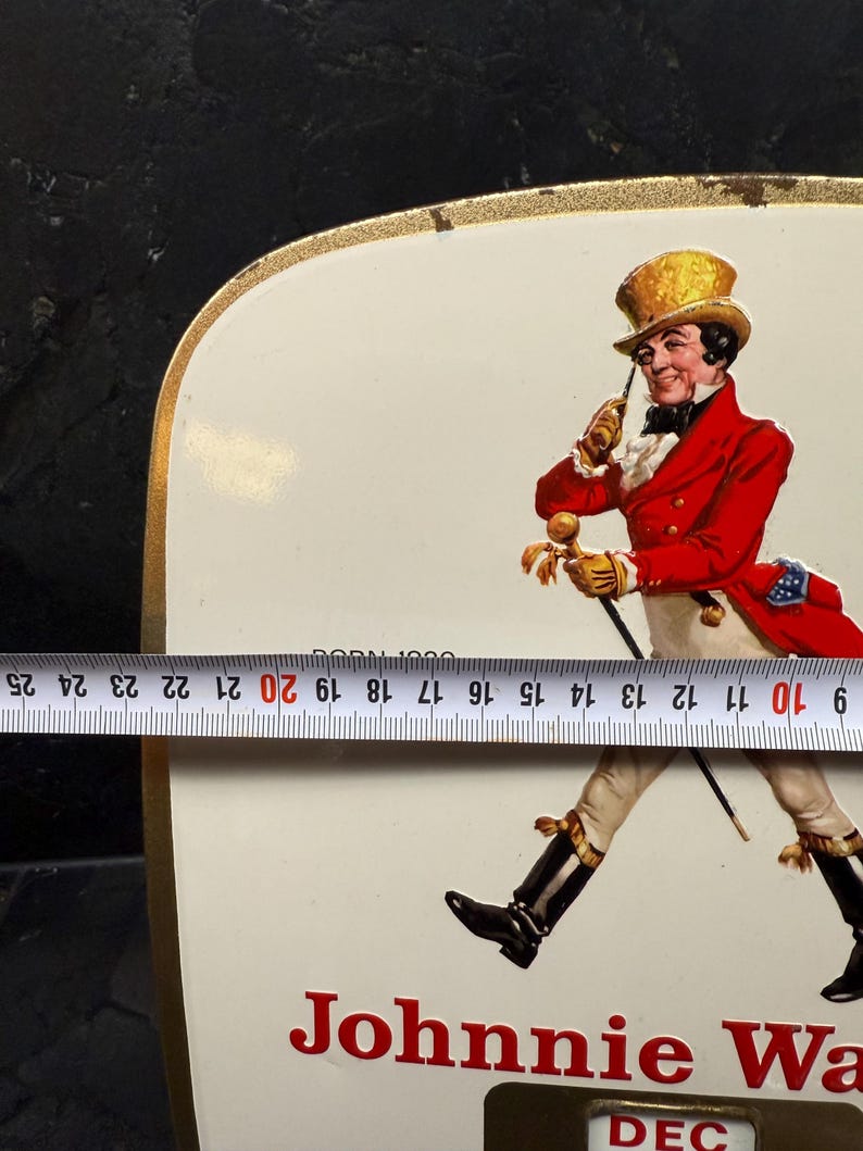 Puede incluir: Letrero met&aacute;lico rectangular vintage con borde dorado. El letrero presenta el logotipo de Johnnie Walker Striding Man en rojo y blanco, con el texto "Johnnie Walker" en la parte inferior. El letrero mide aproximadamente 23 cm de ancho.